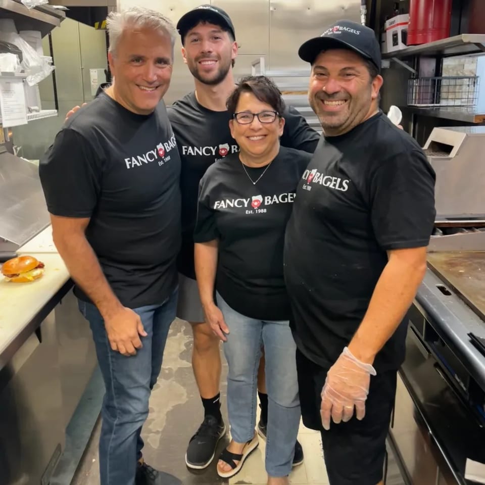 Four smiling Fancy Bagels staff members in black shirts and hats pose in the kitchen.