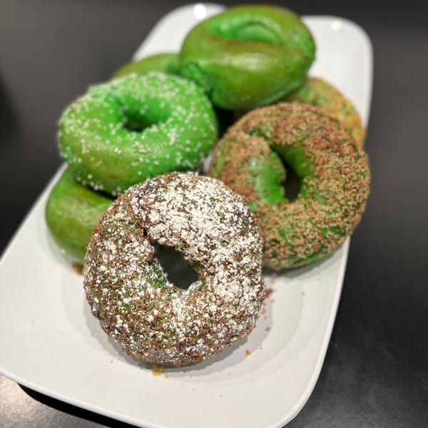 A white platter holds several green bagels, some with white sprinkles, others with brown crumbs and powdered sugar.