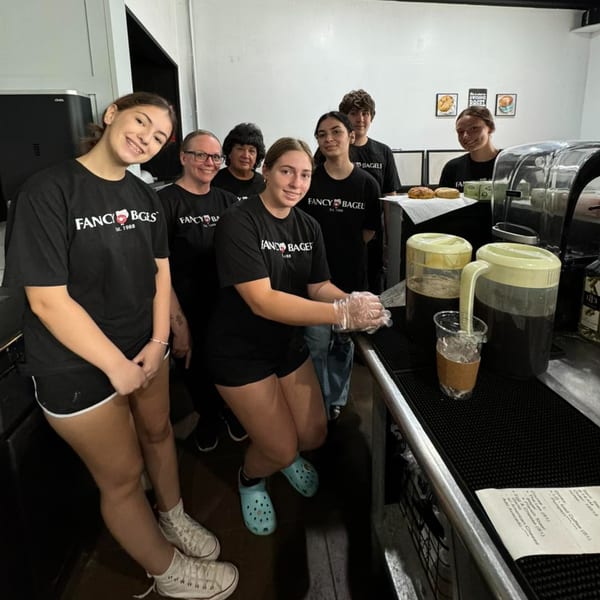Fancy Bagels staff, wearing black t-shirts, smiling behind the counter with drinks and bagels.