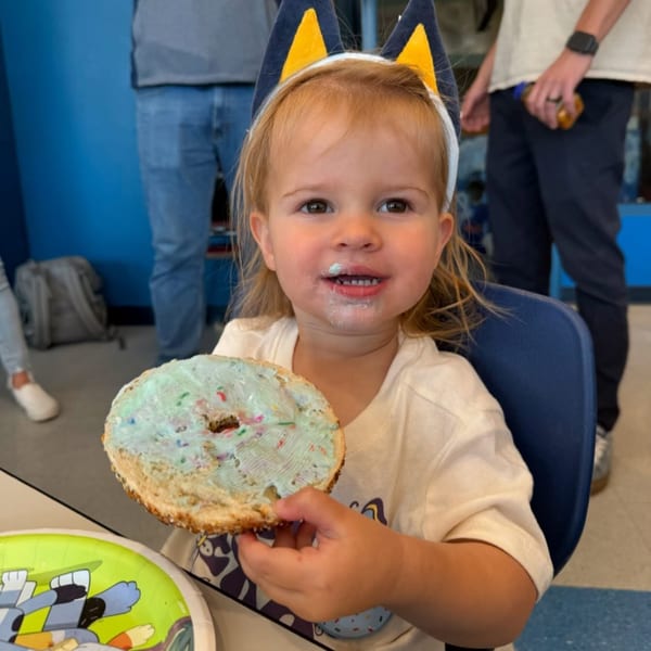 A young child with a blue and yellow headband smiles, holding a bagel with blue cream cheese.