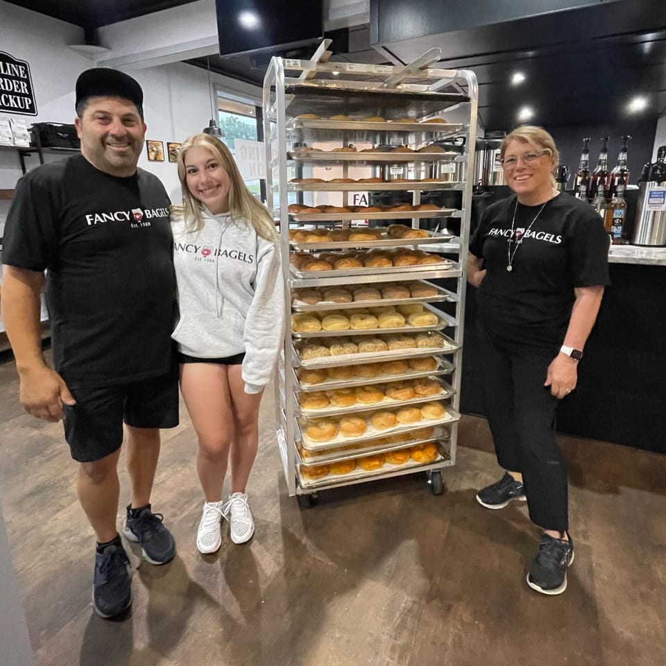 Three Fancy Bagels staff members stand proudly beside a rack filled with freshly baked bagels.