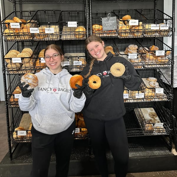 Two smiling Fancy Bagels employees hold bagels in front of a display rack filled with various bagels.