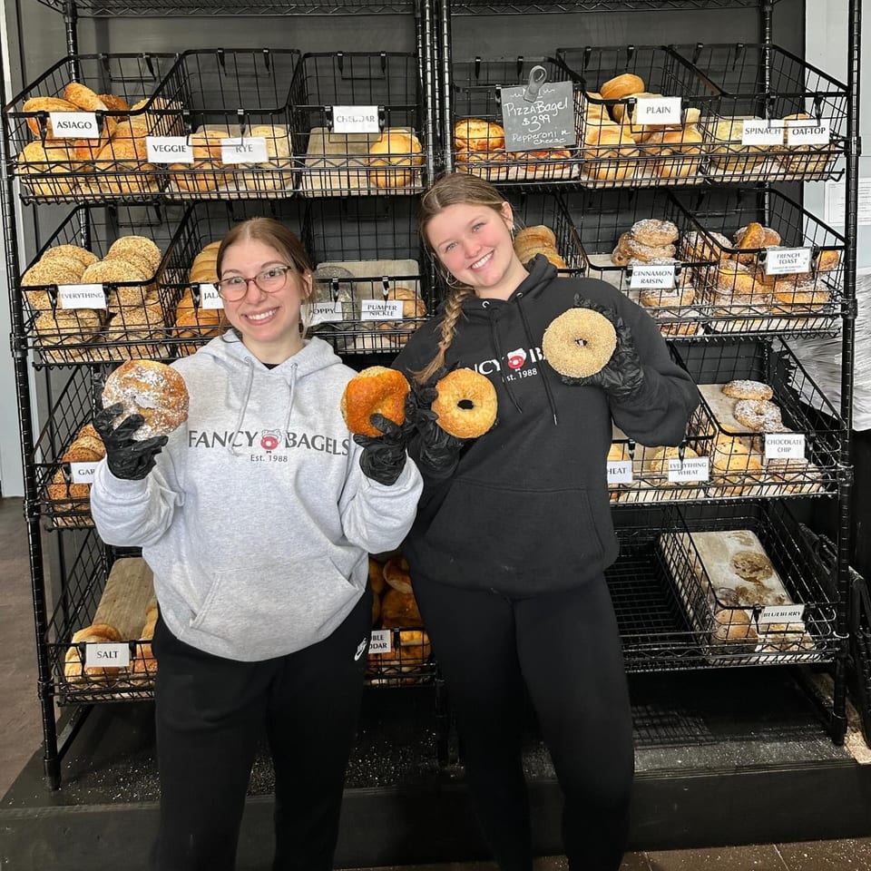 Two smiling Fancy Bagels employees hold bagels in front of a display rack filled with various bagels.