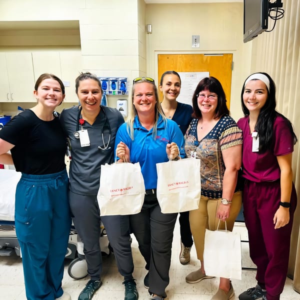Six smiling women, some in scrubs, holding Fancy Bagels bags in a hospital room.