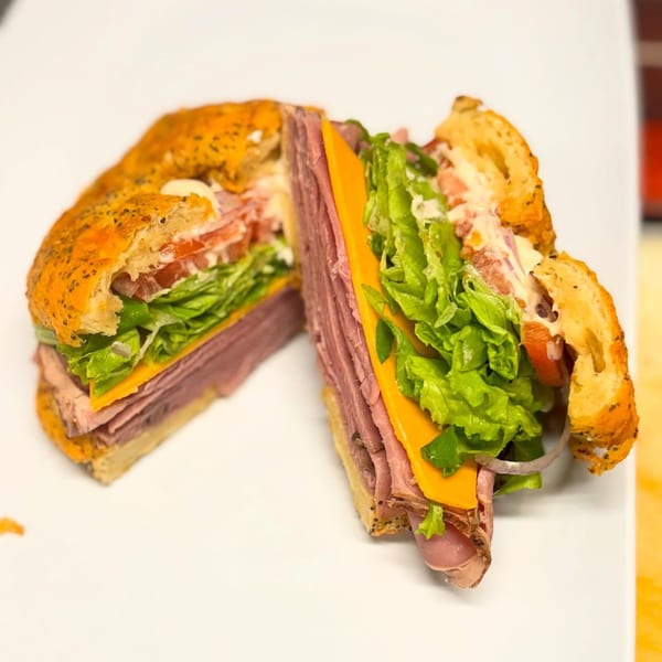 A sliced Fancy Bagels sandwich with roast beef, cheese, lettuce, tomato, and sauce on a white plate.