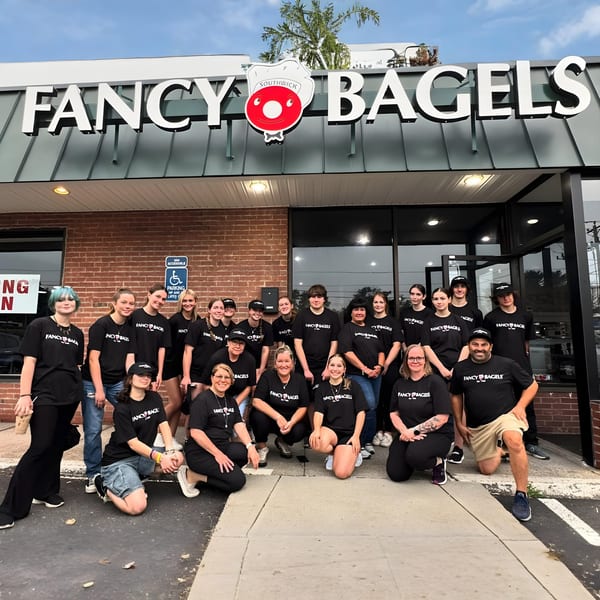 Fancy Bagels staff in black t-shirts pose outside the restaurant under its sign.